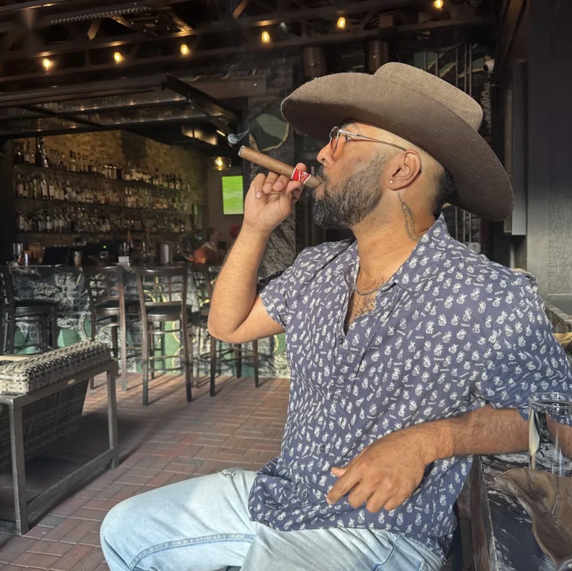 Vighnesh in a cowboy hat and patterned shirt, smoking a cigar at a bar