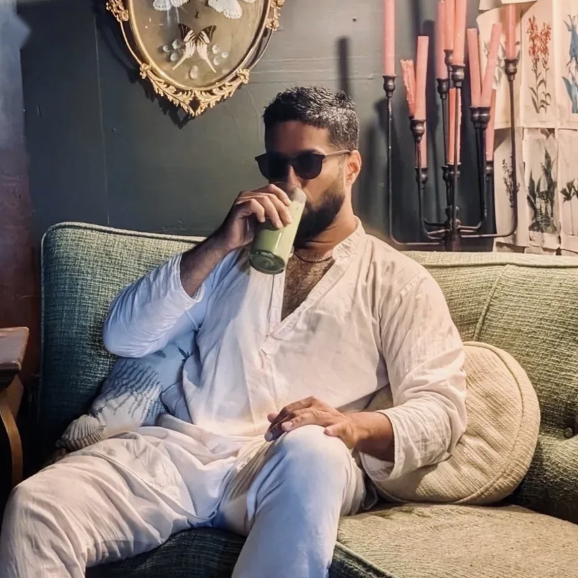 Vighnesh in a white kurta and sunglasses on a green sofa, sipping a drink