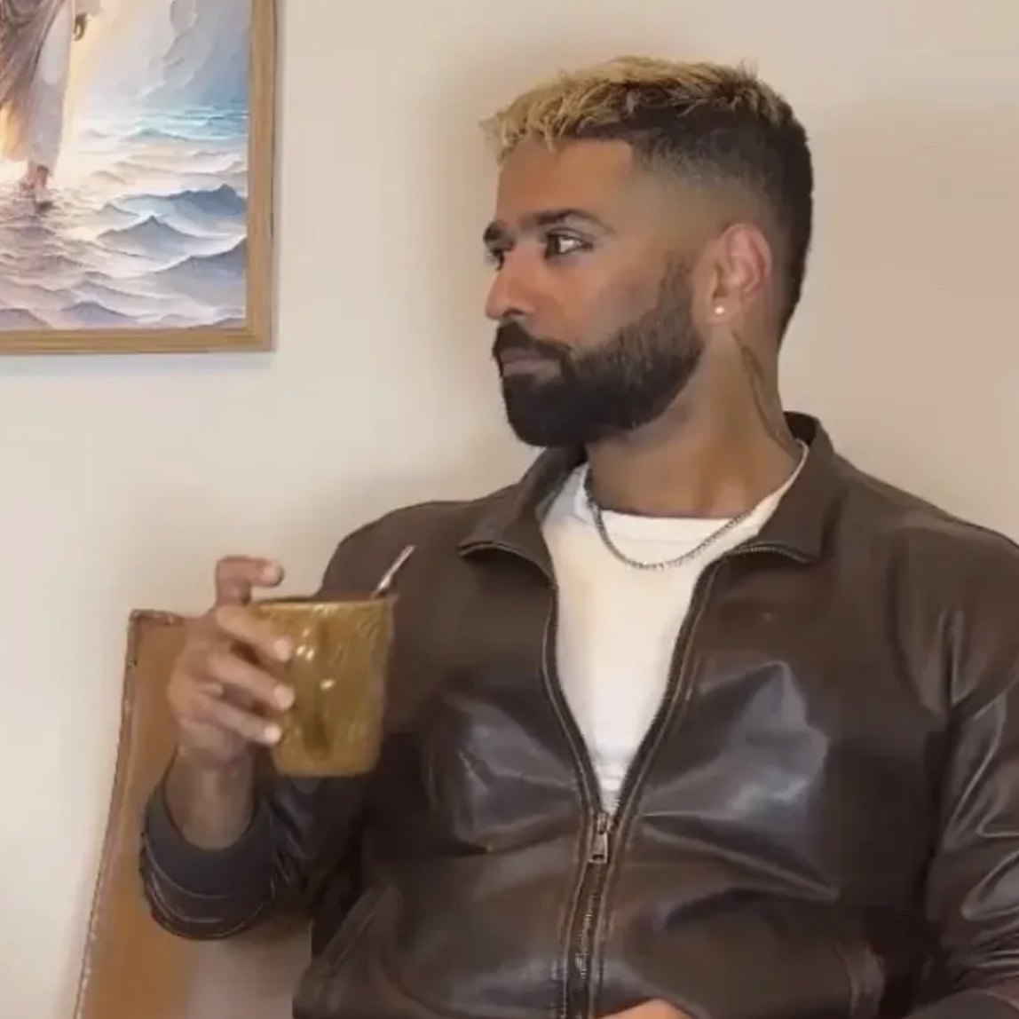 Vighnesh in a brown leather jacket holding a glass, beside a painting of Jesus walking on water
