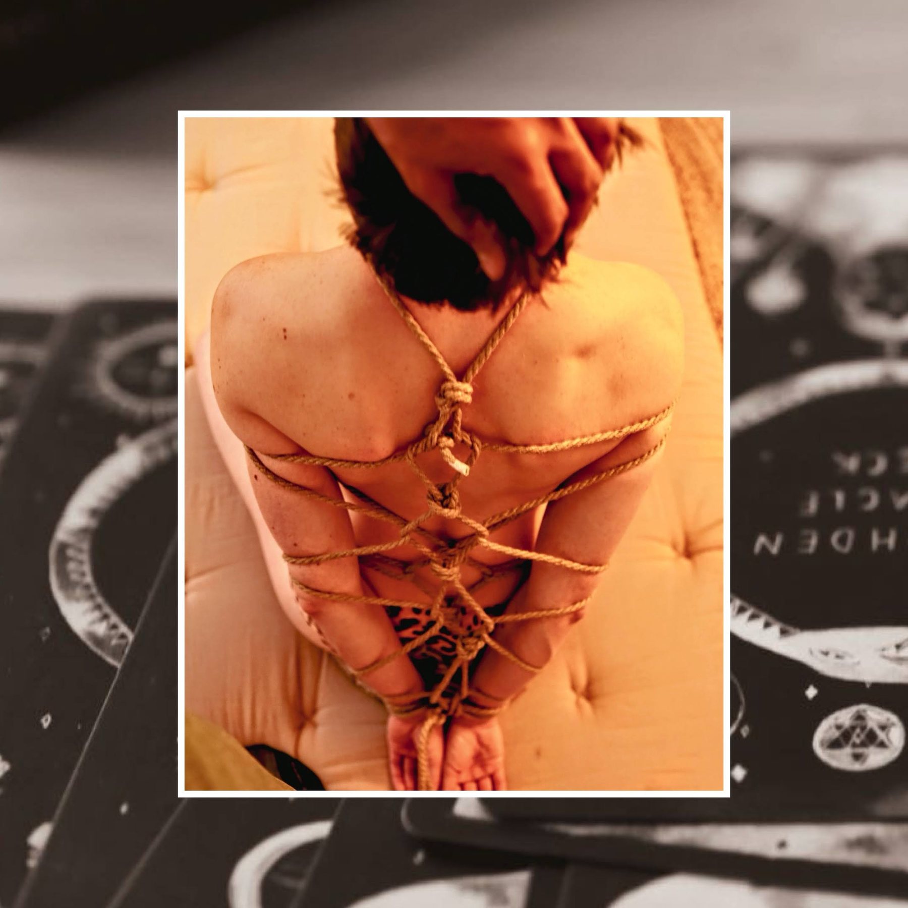 A kneeling figure seen from above in a rope harness, arms tied behind, framed over a deck of cards